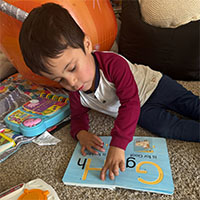 A young child with dark hair lies on the carpet, tracing letters in a book. The image is mostly black and white, but the child’s long-sleeve shirt stands out in bright red. Toys and pillows are nearby.