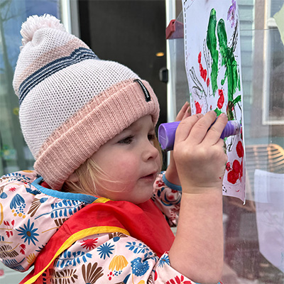 A young child in a pink and white knit hat colors a drawing with a purple marker, wearing a colorful jacket and a red apron, focused on the artwork taped to a window.