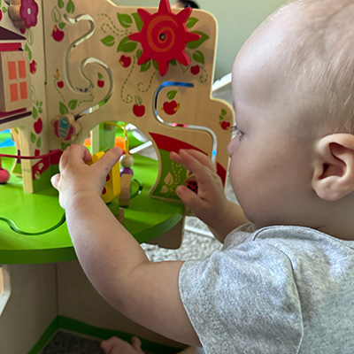 A baby in a gray shirt plays with a colorful wooden activity center, moving beads along a wire maze. The focus is on the childs hands and the interactive toy.